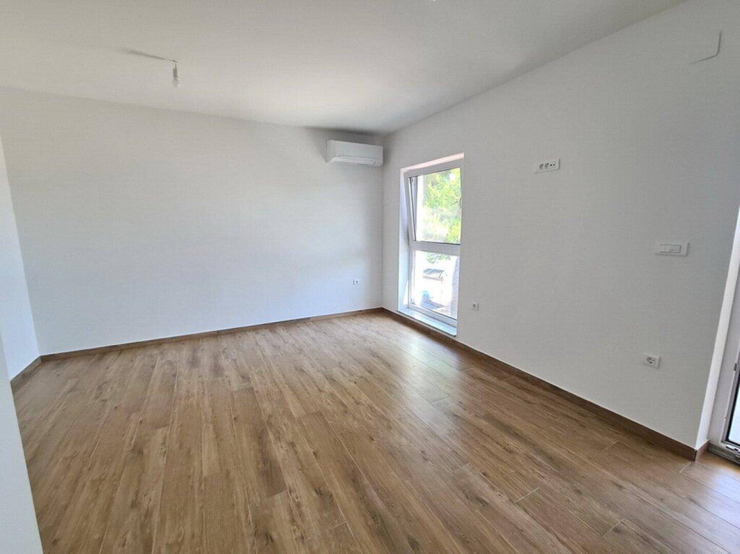 Helles, leeres Zimmer mit Holzboden, weißen Wänden und installierter Klimaanlage.
