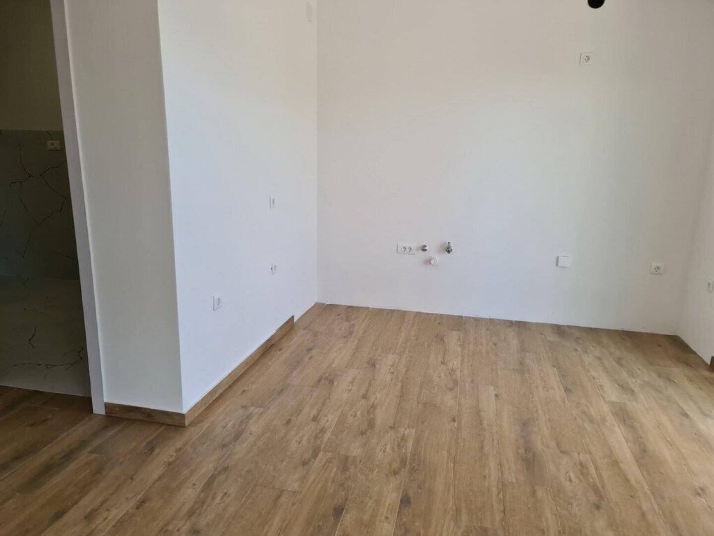 Leerer Raum mit Holzboden und sichtbaren Anschlüssen an der Wand, möglicherweise für eine Küche.