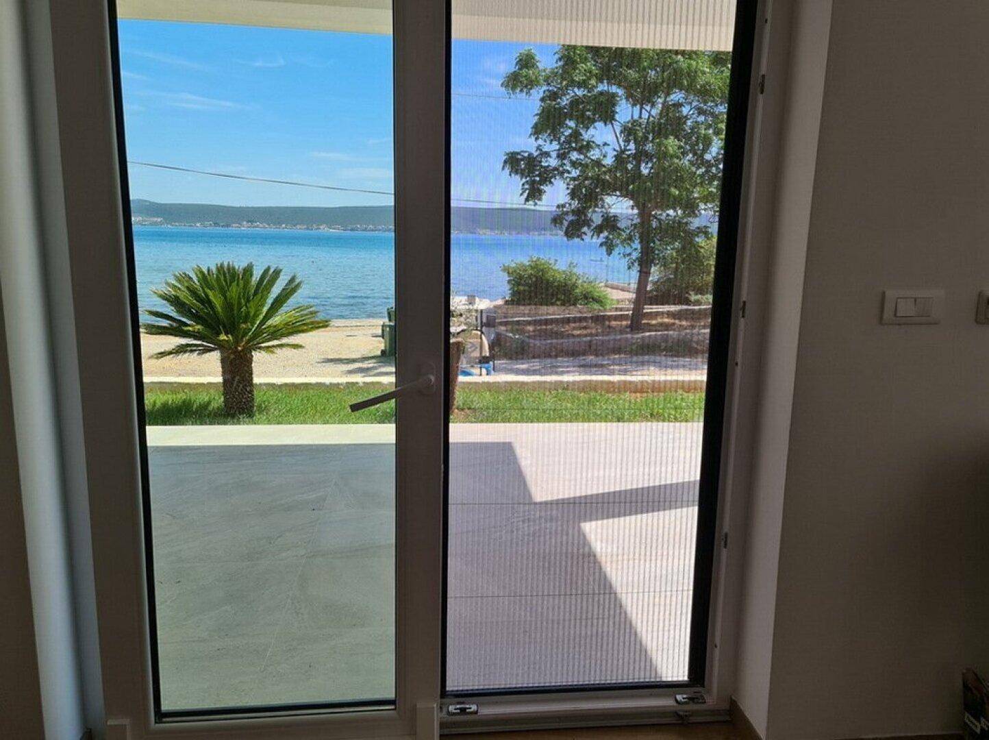 Blick aus dem Zimmer durch eine große Tür auf das blaue Meer und die grüne Küstenlandschaft.