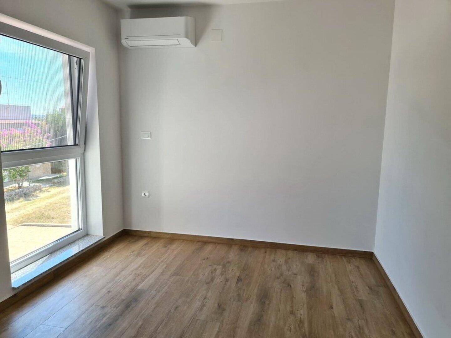 Helles, leeres Zimmer mit Holzboden, großem Fenster und installierter Klimaanlage.