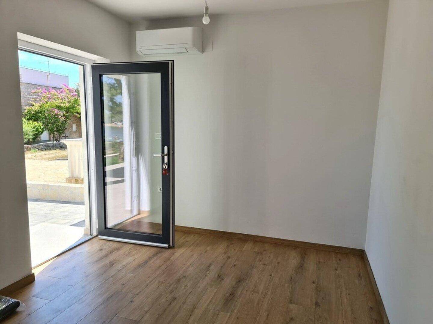 Helles, leeres Zimmer mit Holzboden und einer Glastür, die zu einem Außenbereich führt.