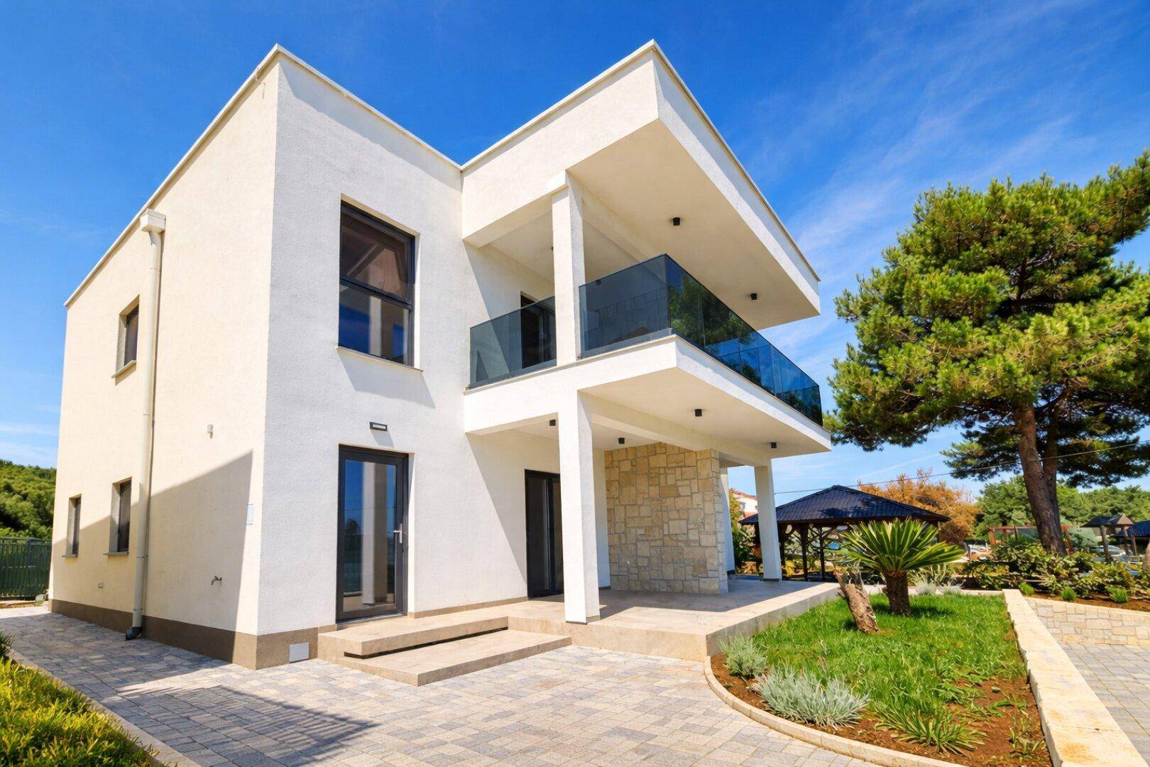 Moderne weiße Villa mit klaren Linien, Glasbalkonen und Steinakzenten unter blauem Himmel.