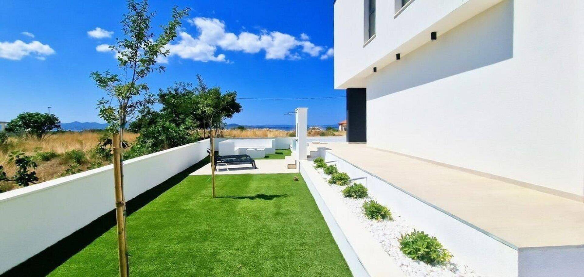 Schmaler Gartenstreifen mit Kunstrasen und kleinen Pflanzen entlang der modernen Hausfassade.
