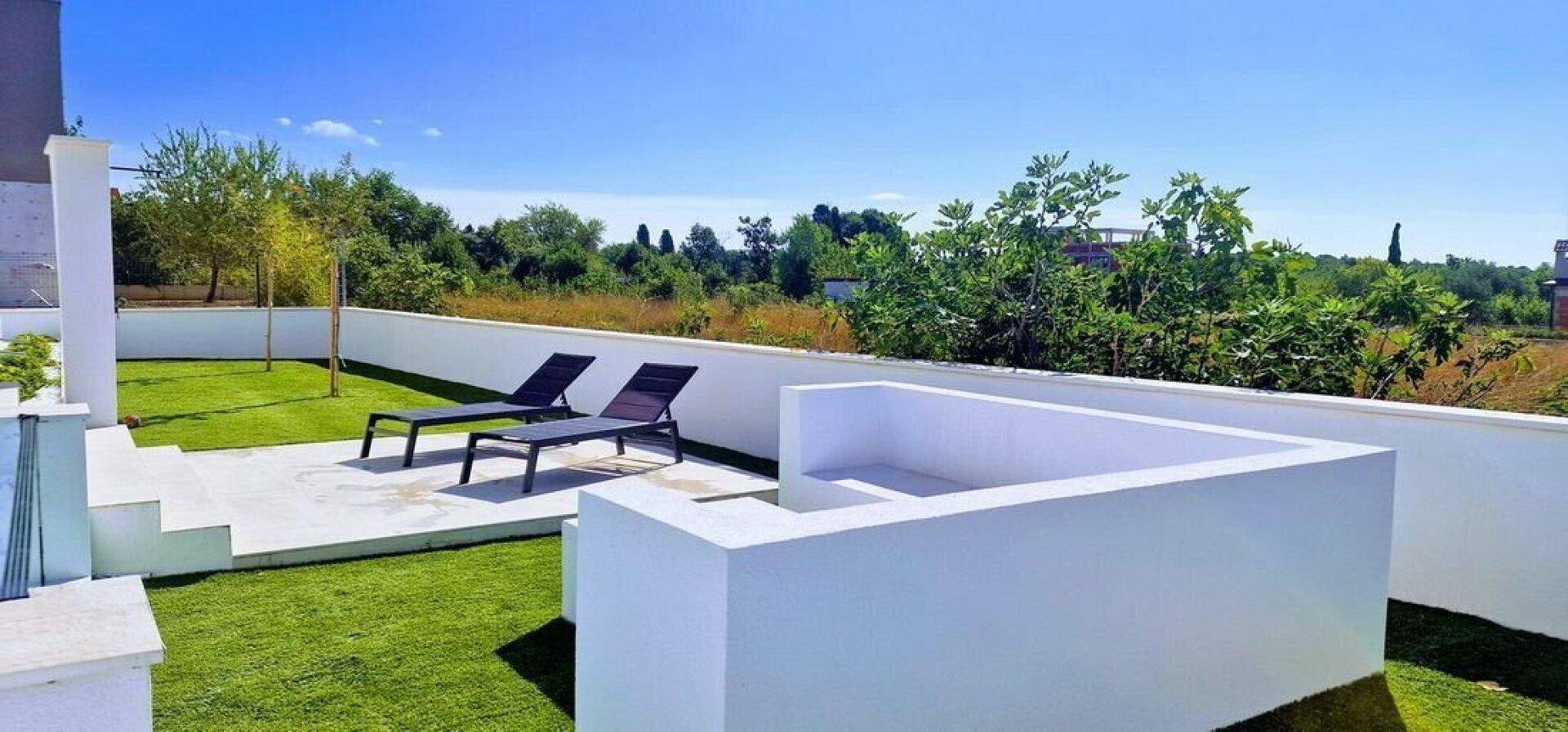 Gepflegter Gartenbereich mit Kunstrasen, Liegestühlen und einer niedrigen weißen Mauer unter blauem Himmel.