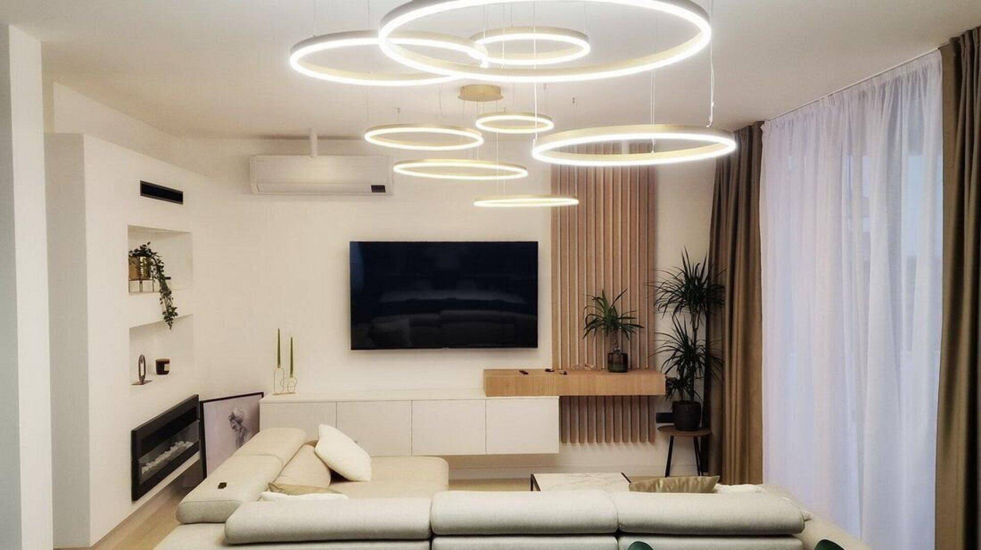 Modernes Wohnzimmer mit bequemer Couch, Wandfernseher und auffälligen Deckenleuchten.