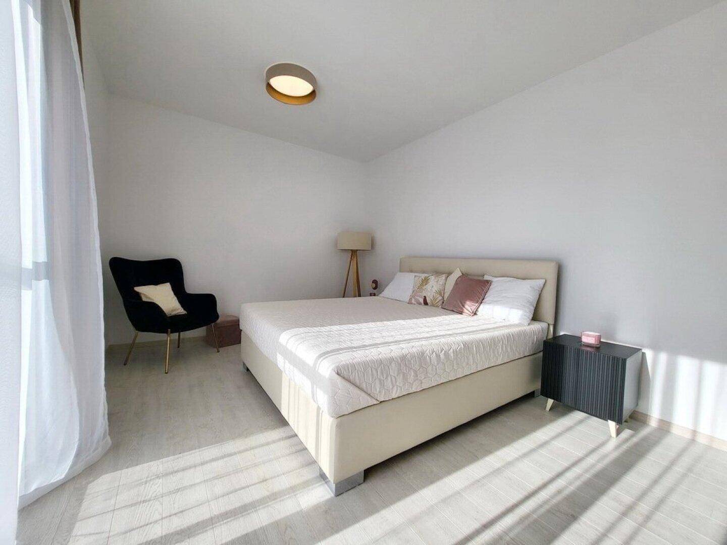 Helles Schlafzimmer mit beigem Bett, Lesesessel und viel natürlichem Licht durch das Fenster.