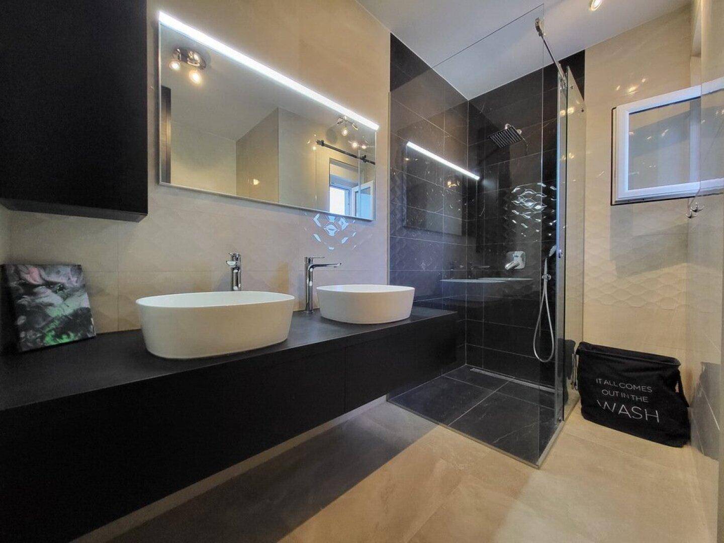 Elegantes Badezimmer mit Doppelwaschbecken, großer begehbarer Dusche und dunklen Fliesen.