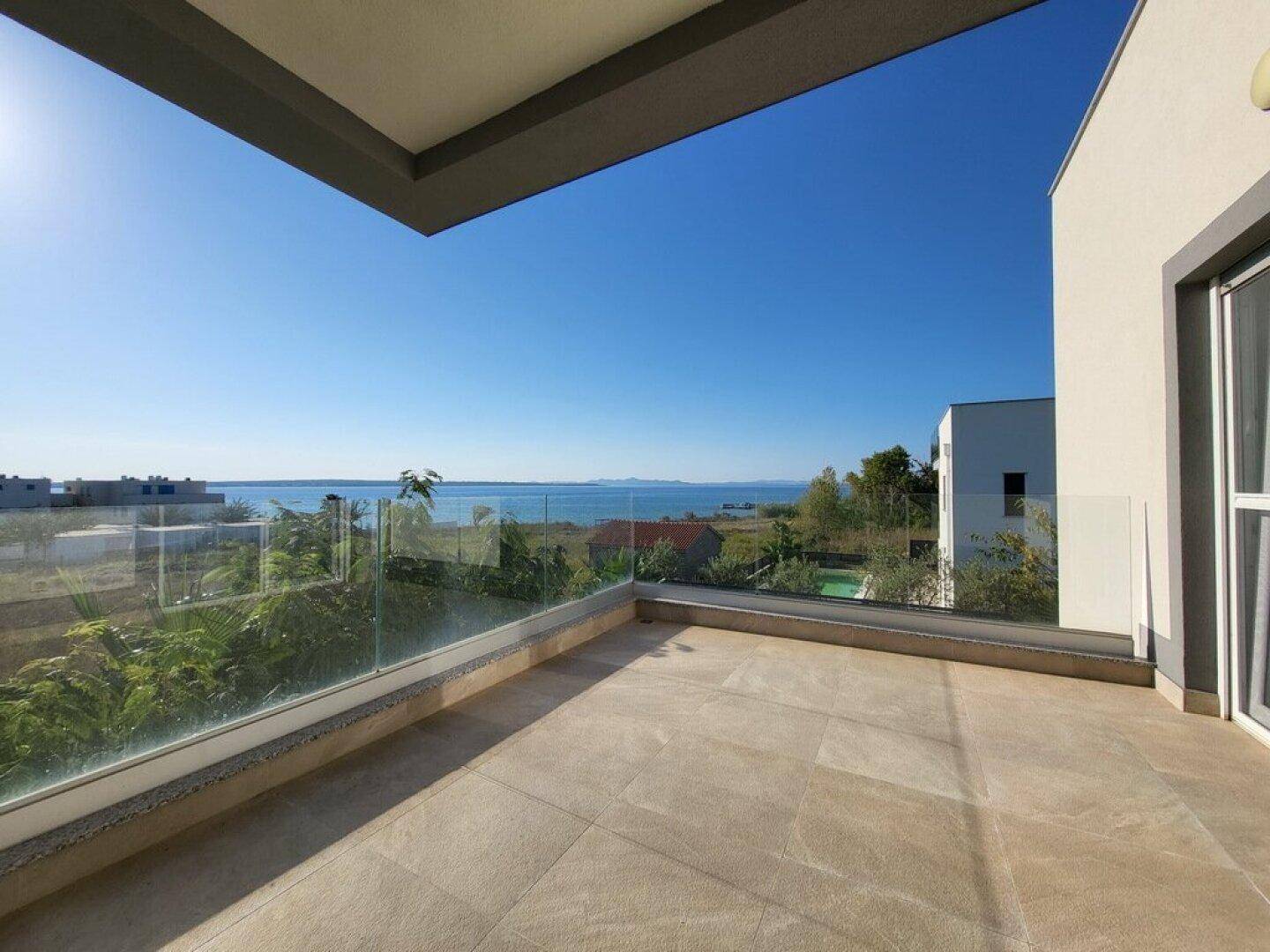 Geräumiger Balkon mit Glasgeländer und weitem Blick auf das Meer unter strahlendem Sonnenschein.
