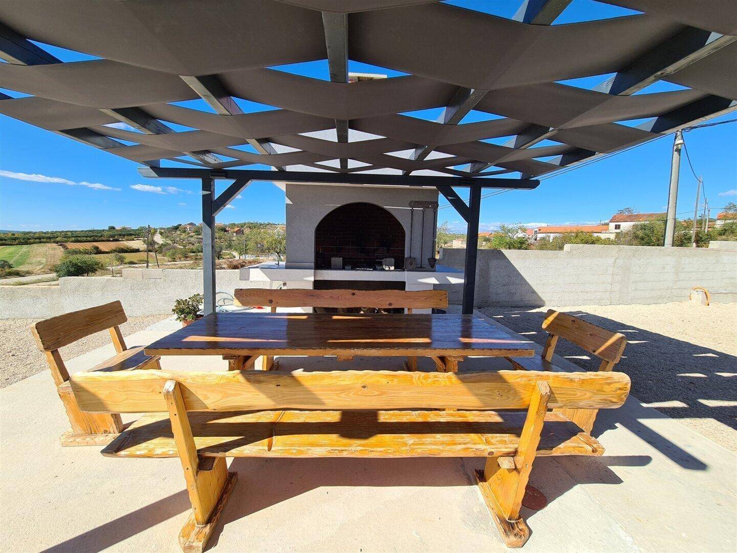 Gemütlicher Essbereich im Freien unter einer Pergola mit großem Holztisch und Grill.