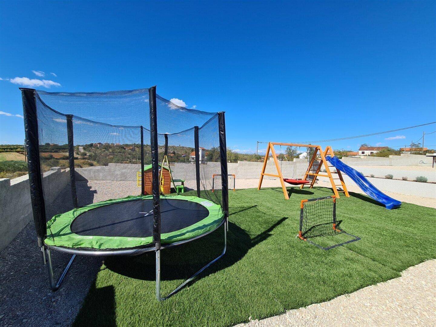 Kinderspielplatz mit Trampolin, Schaukel und Rutsche auf grünem Kunstrasen.