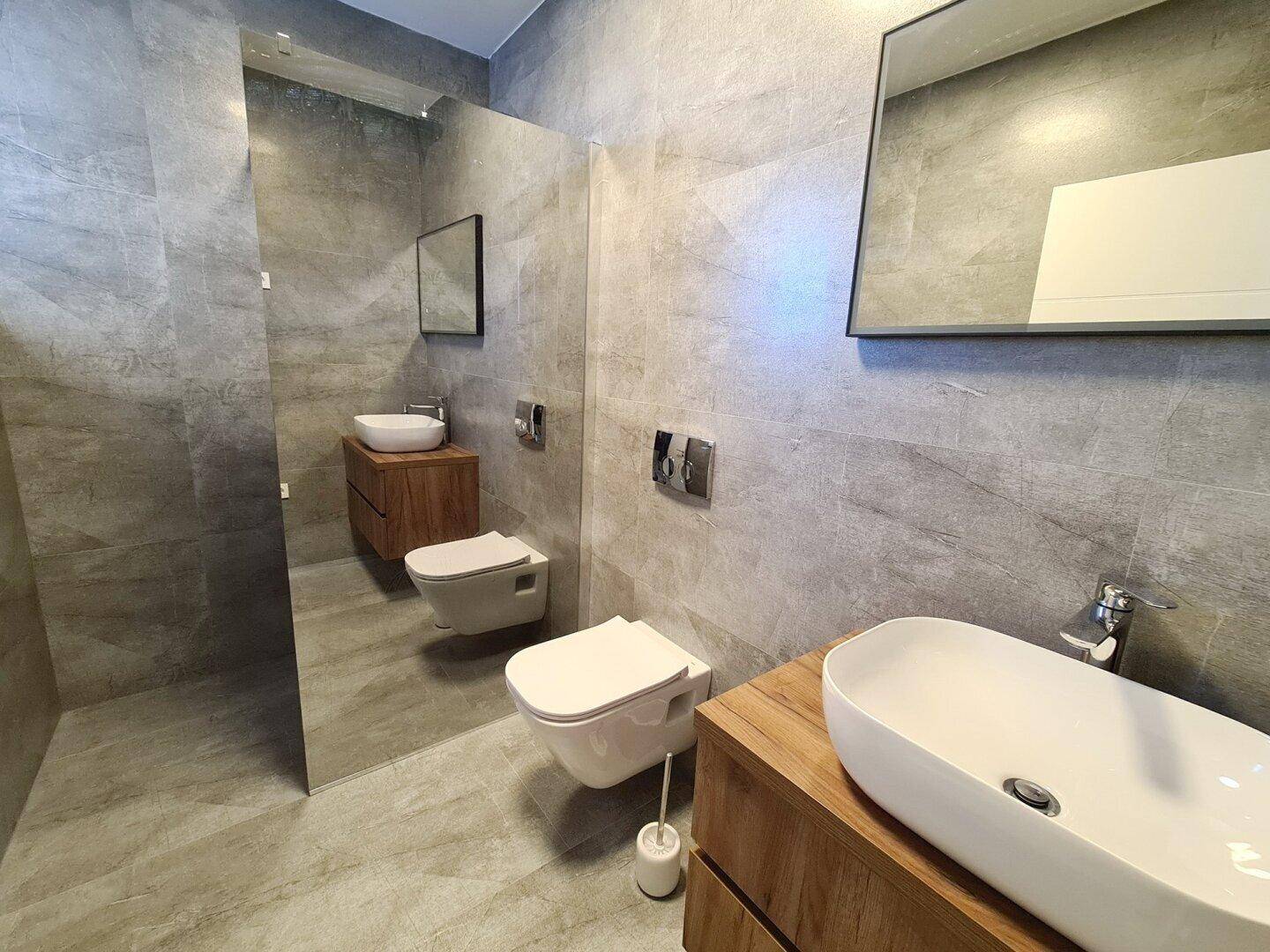 Modernes Badezimmer mit grauen Fliesen, zwei Waschbecken und wandhängenden Toiletten.