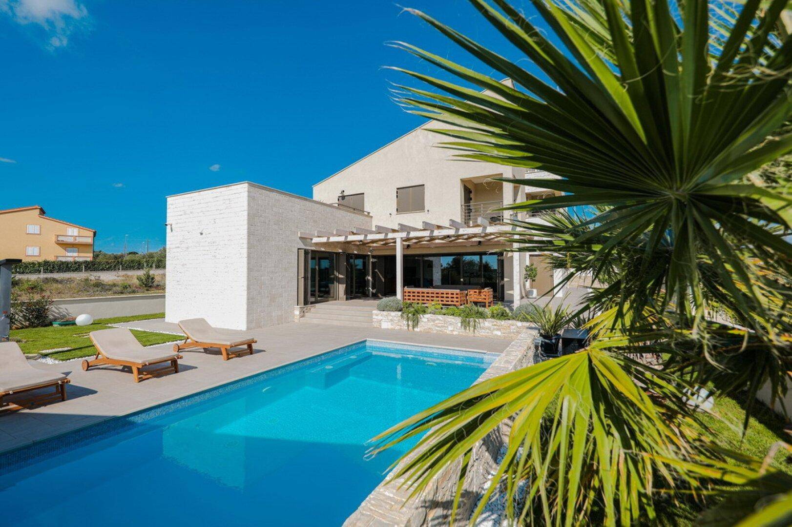 Moderne Villa mit großem Pool, Liegestühlen und Palmen unter blauem Himmel.
