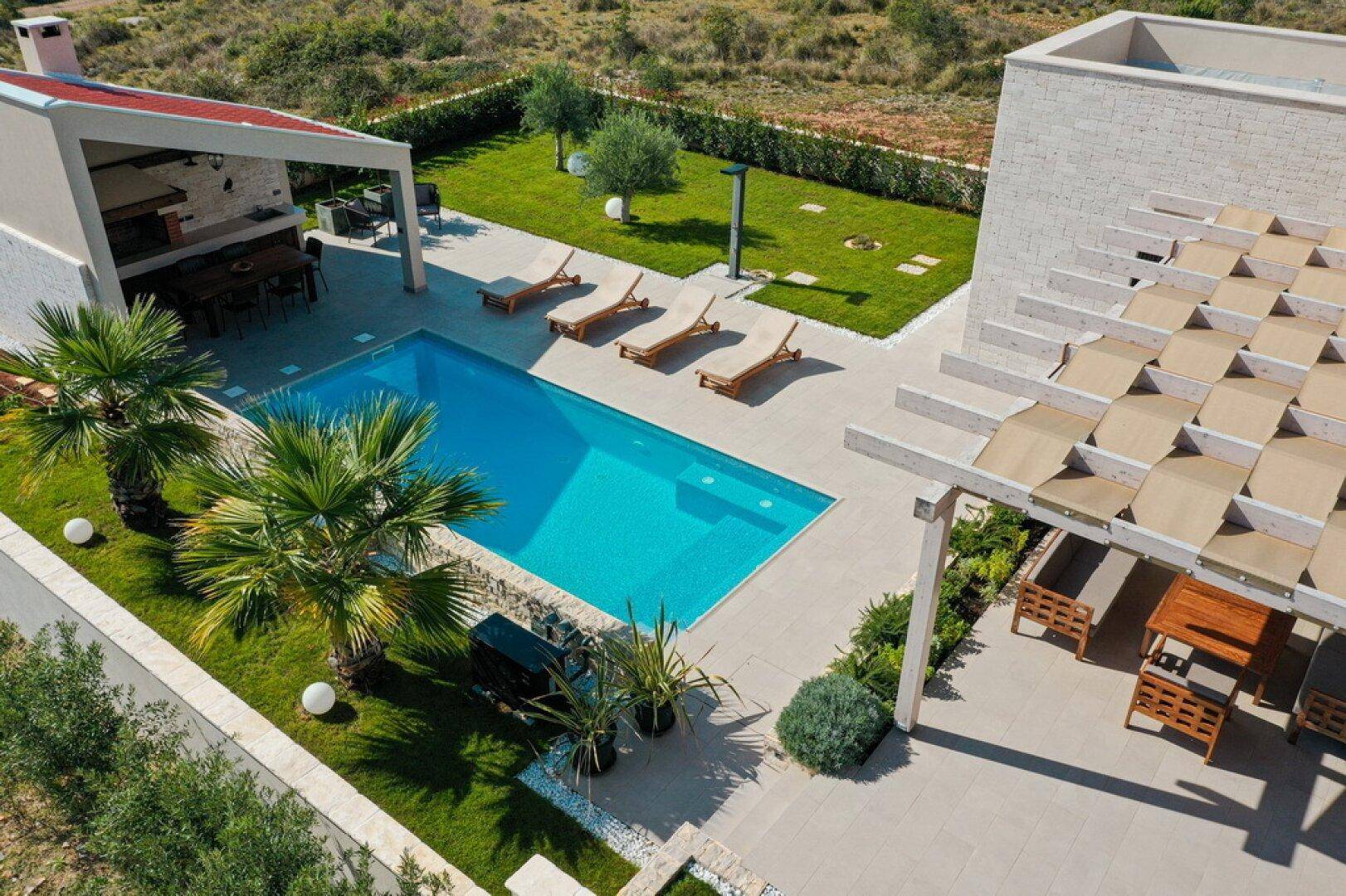 Ansicht von oben auf den Poolbereich mit Palmen, Liegestühlen und überdachter Terrasse.