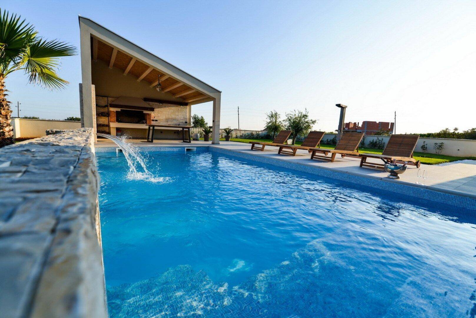 Erfrischender Pool mit Wasserspiel und überdachtem Grillbereich, umgeben von Sonnenliegen.