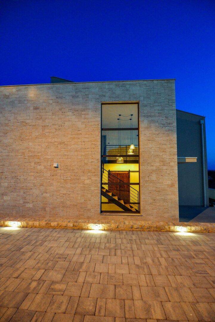 Moderne Steinfassade der Villa mit großem Fenster und beleuchtetem Eingangsbereich bei Nacht.