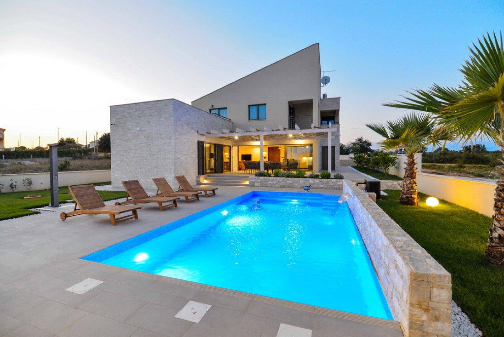 Elegante Villa mit beleuchtetem Pool und Palmen im Garten bei Sonnenuntergang.