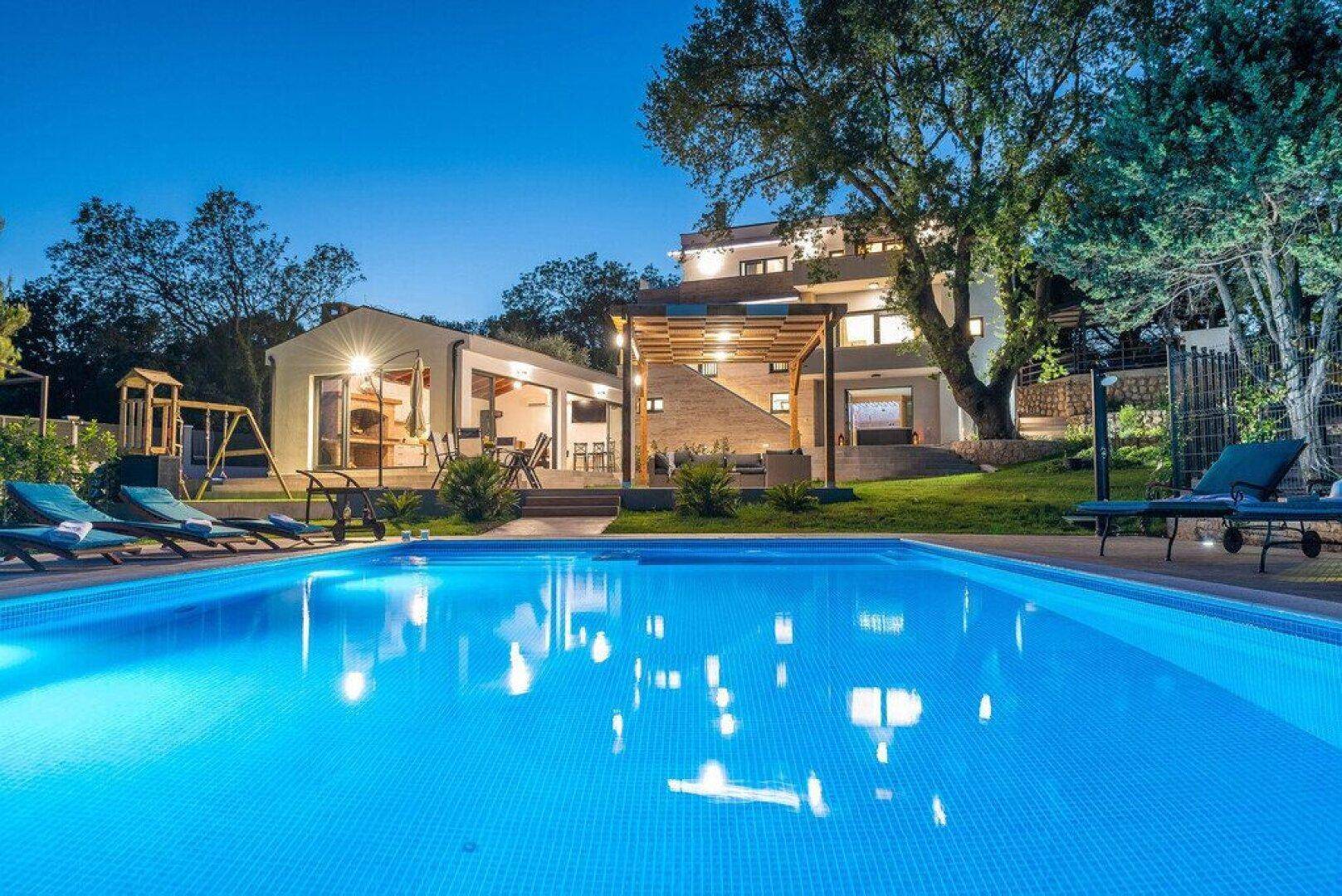 Beleuchteter Poolbereich bei Nacht mit Garten und Spielplatz.