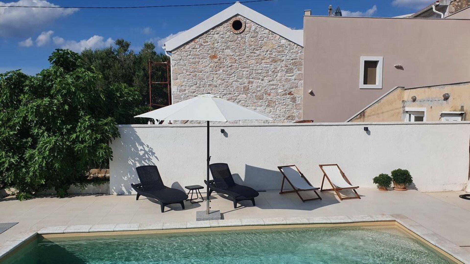 Außenbereich mit Pool, Liegestühlen und Sonnenschirm vor einem Steinhaus.