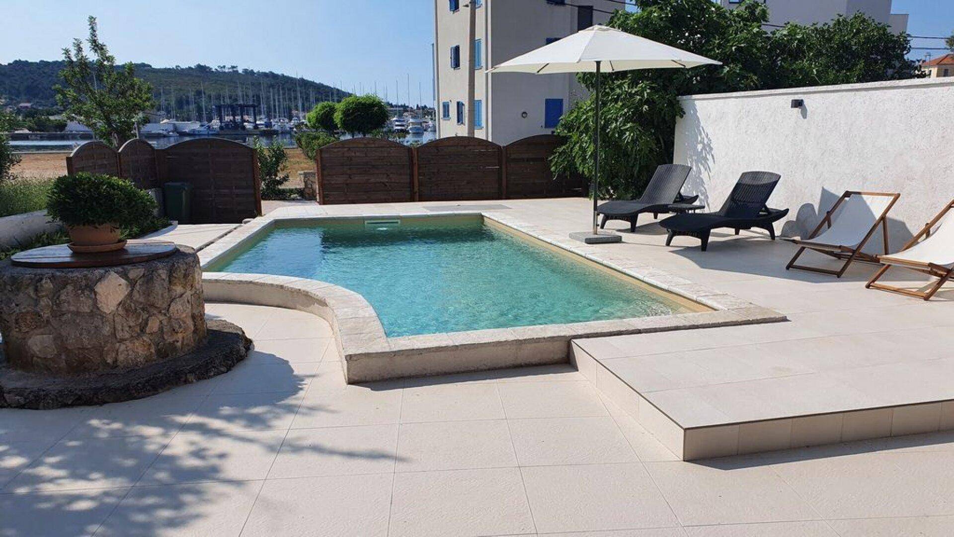 Poolbereich mit Liegestühlen und Sonnenschirm, mit Blick auf den Hafen und das Meer.
