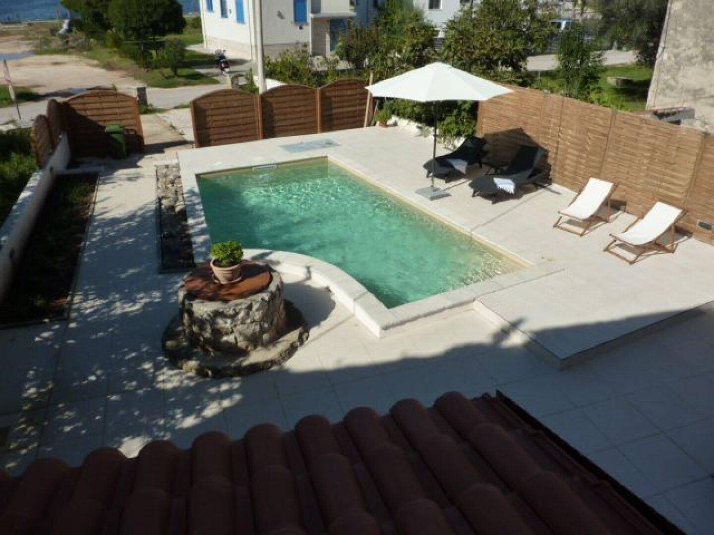 Vogelperspektive des Poolbereichs mit Liegestühlen, Sonnenschirm und Holzzaun.
