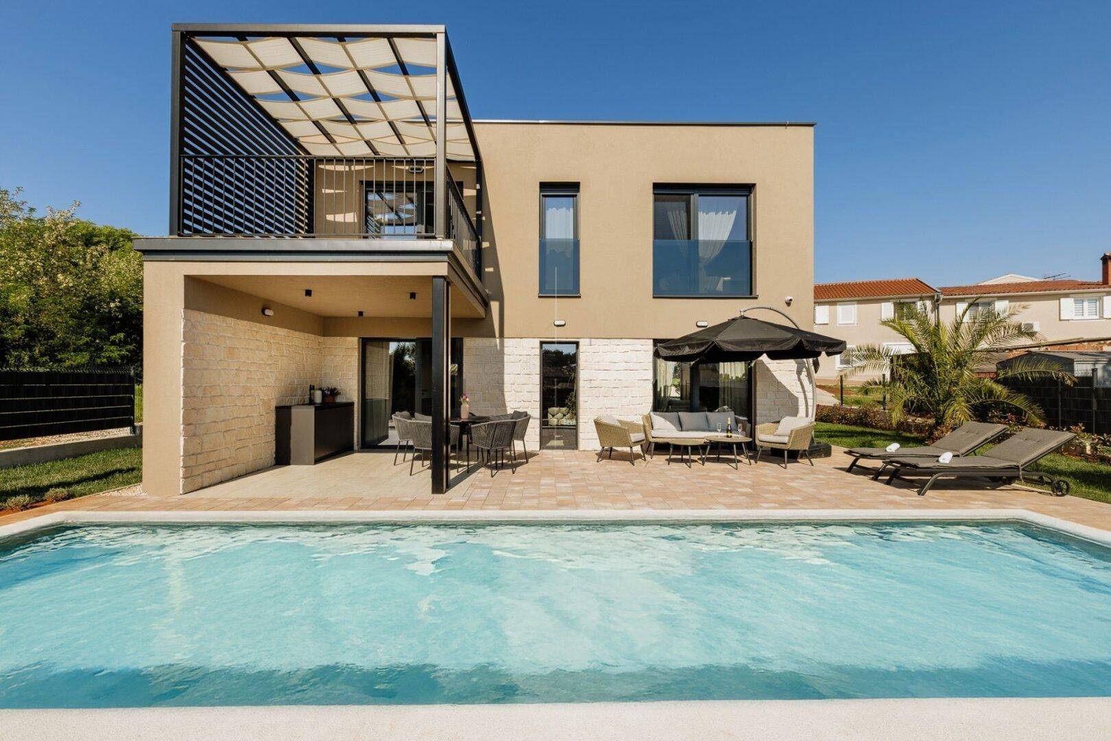 Ansicht der modernen Villa mit Poolbereich, Terrasse und überdachtem Balkon unter blauem Himmel.