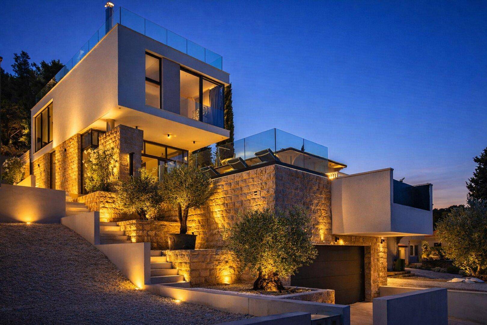 Moderne Villa bei Nacht mit beleuchteten Steinfassaden und Terrassenbereichen.