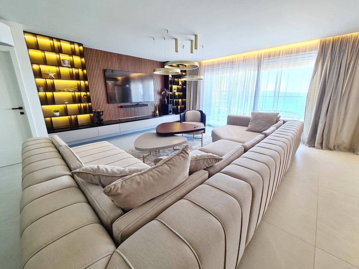 Helles Wohnzimmer mit beigem Ecksofa, modernen Leuchten und raumhohen Fenstern mit Meerblick.