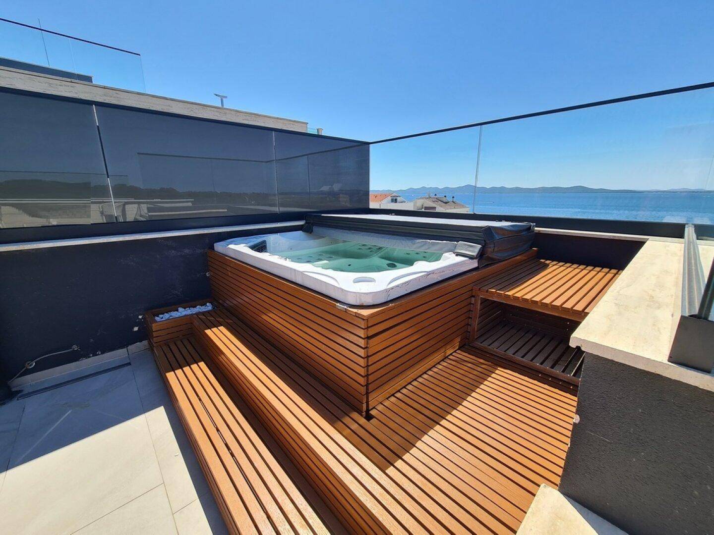 Dachterrasse mit Whirlpool, Holzdeck und Panoramablick auf das Meer unter blauem Himmel.