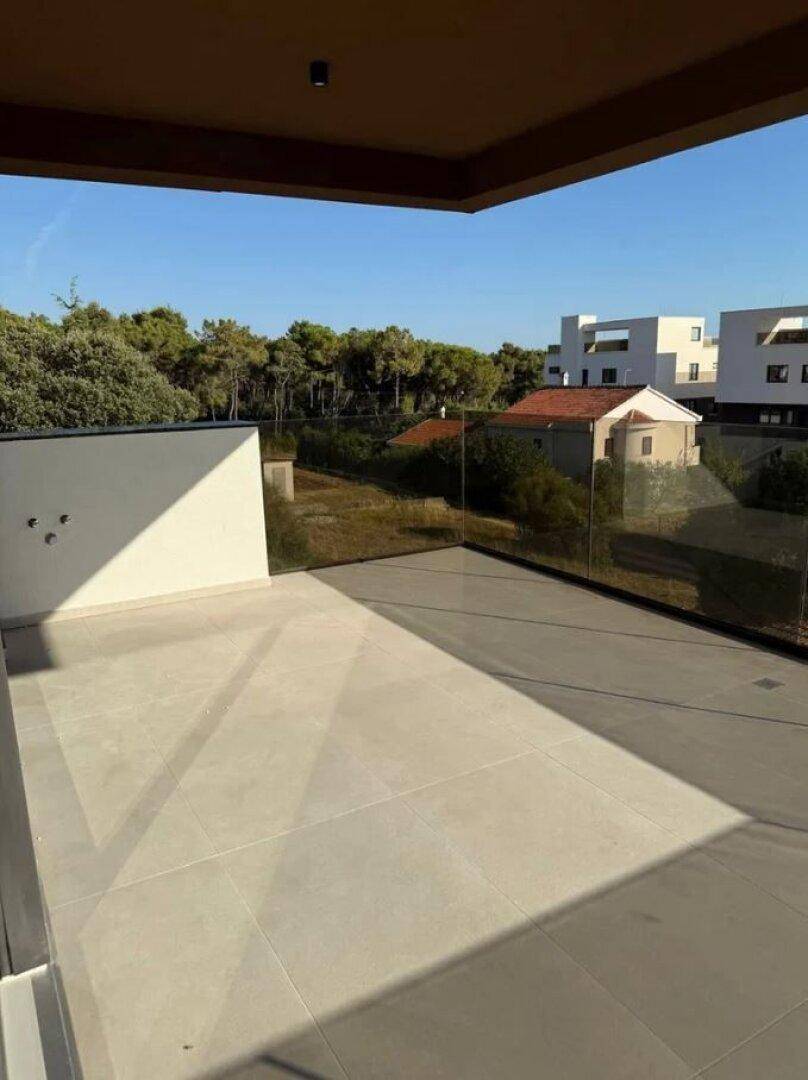 Große, leere Terrasse mit Glasgeländer und Blick auf die grüne Umgebung und Nachbargebäude.