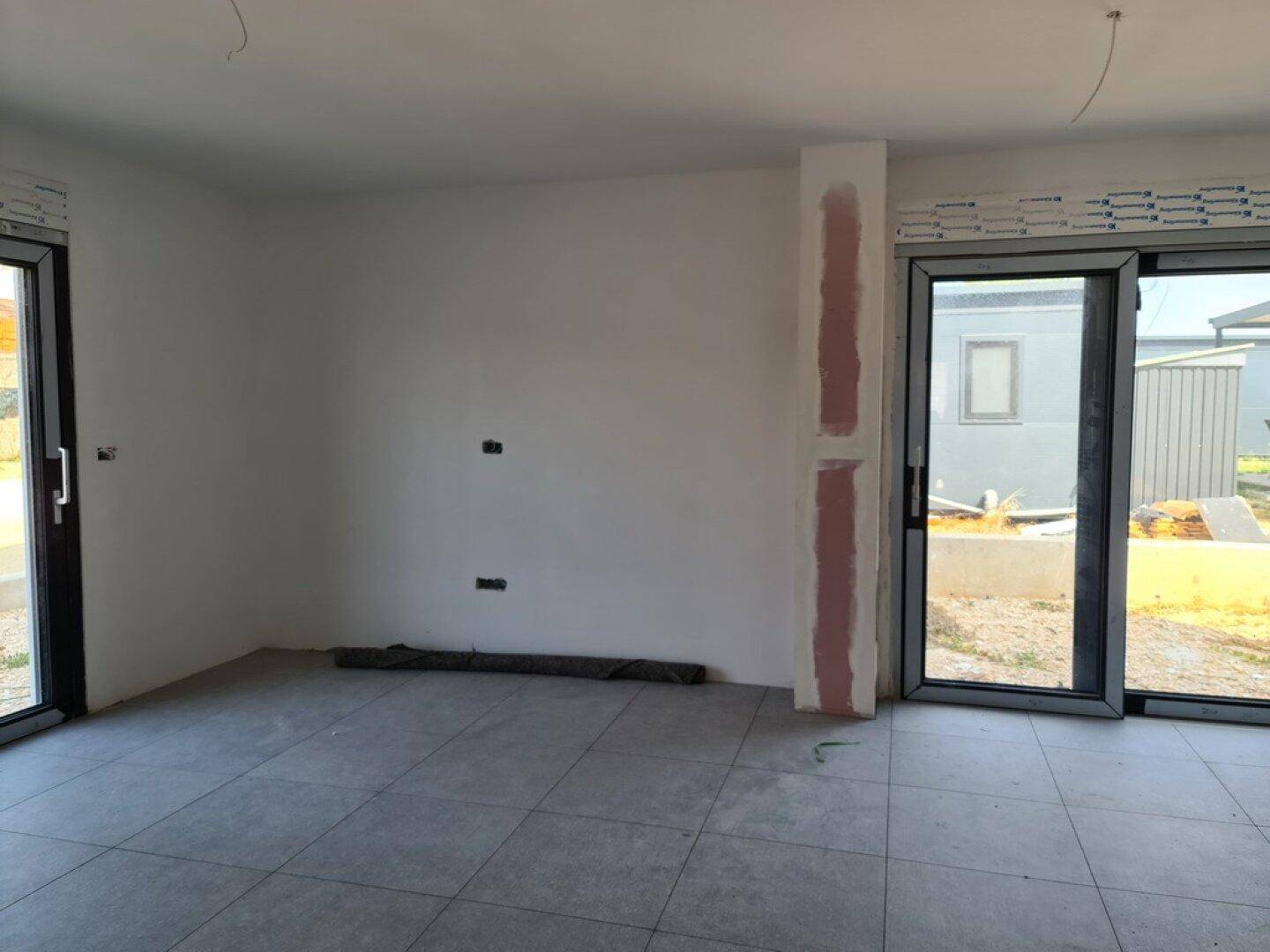 Unfertiger Innenraum mit grauen Fliesen, weißen Wänden und großen Fenstertüren.