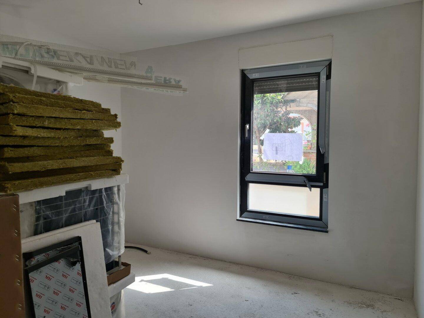 Unfertiges Zimmer mit weiß gestrichenen Wänden und einem einzelnen Fenster, Baumaterialien sichtbar.