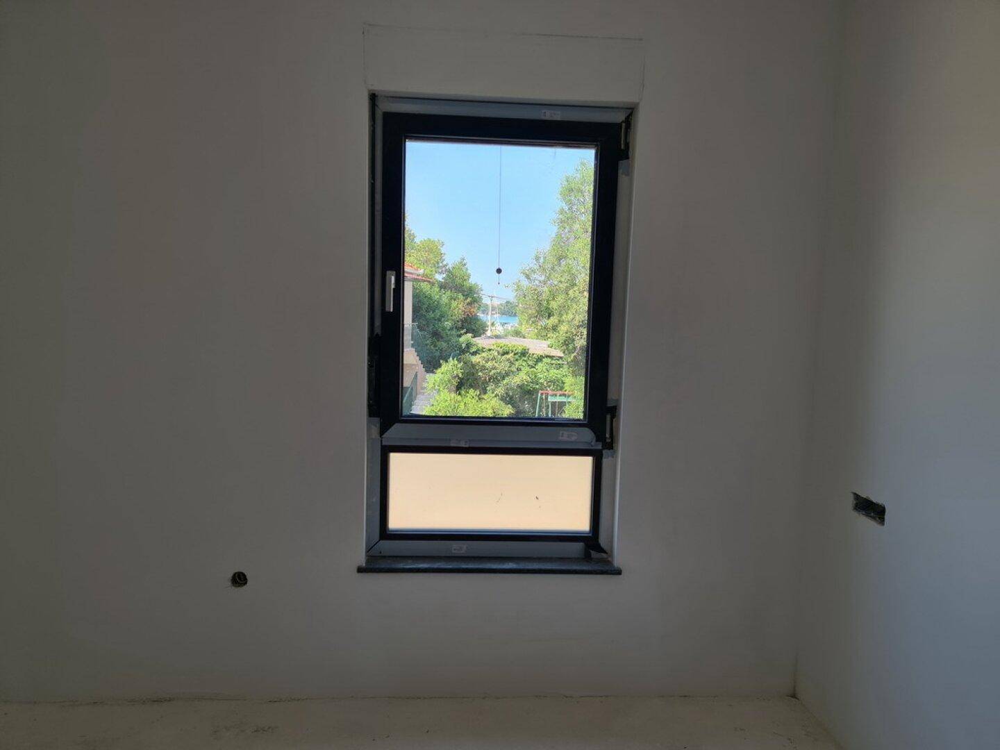 Unfertiges Zimmer mit weiß gestrichenen Wänden und einem Fenster mit Blick auf Bäume.