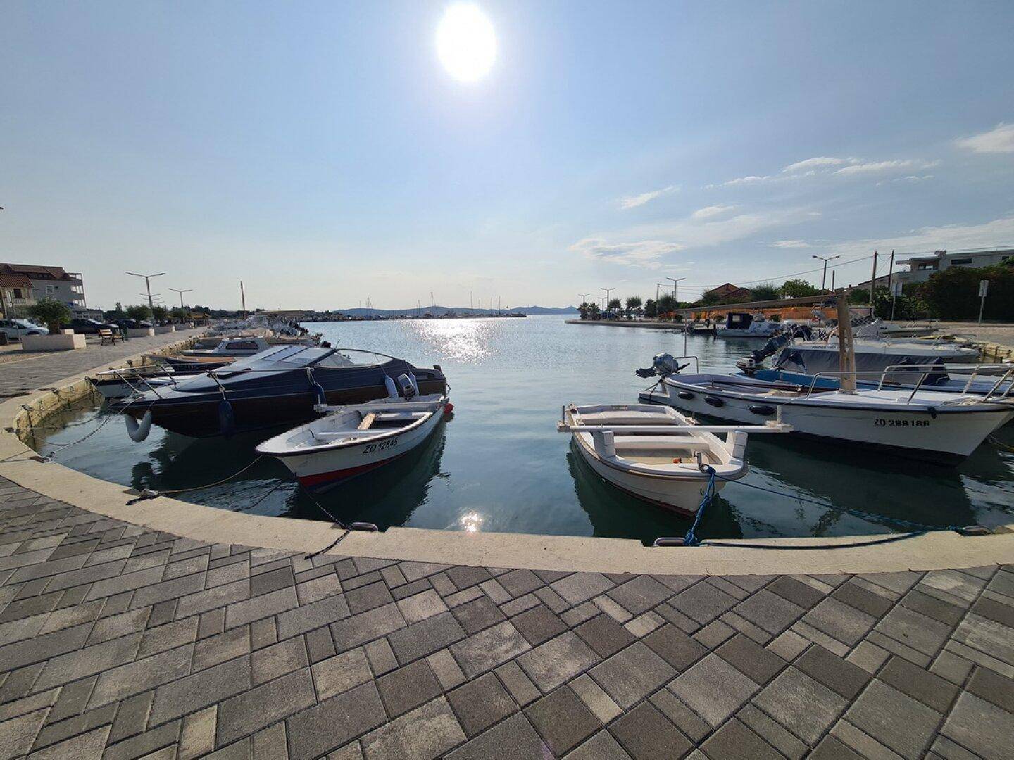 Sonniger Blick auf einen belebten Hafen mit vielen Booten und einer gepflasterten Uferpromenade.