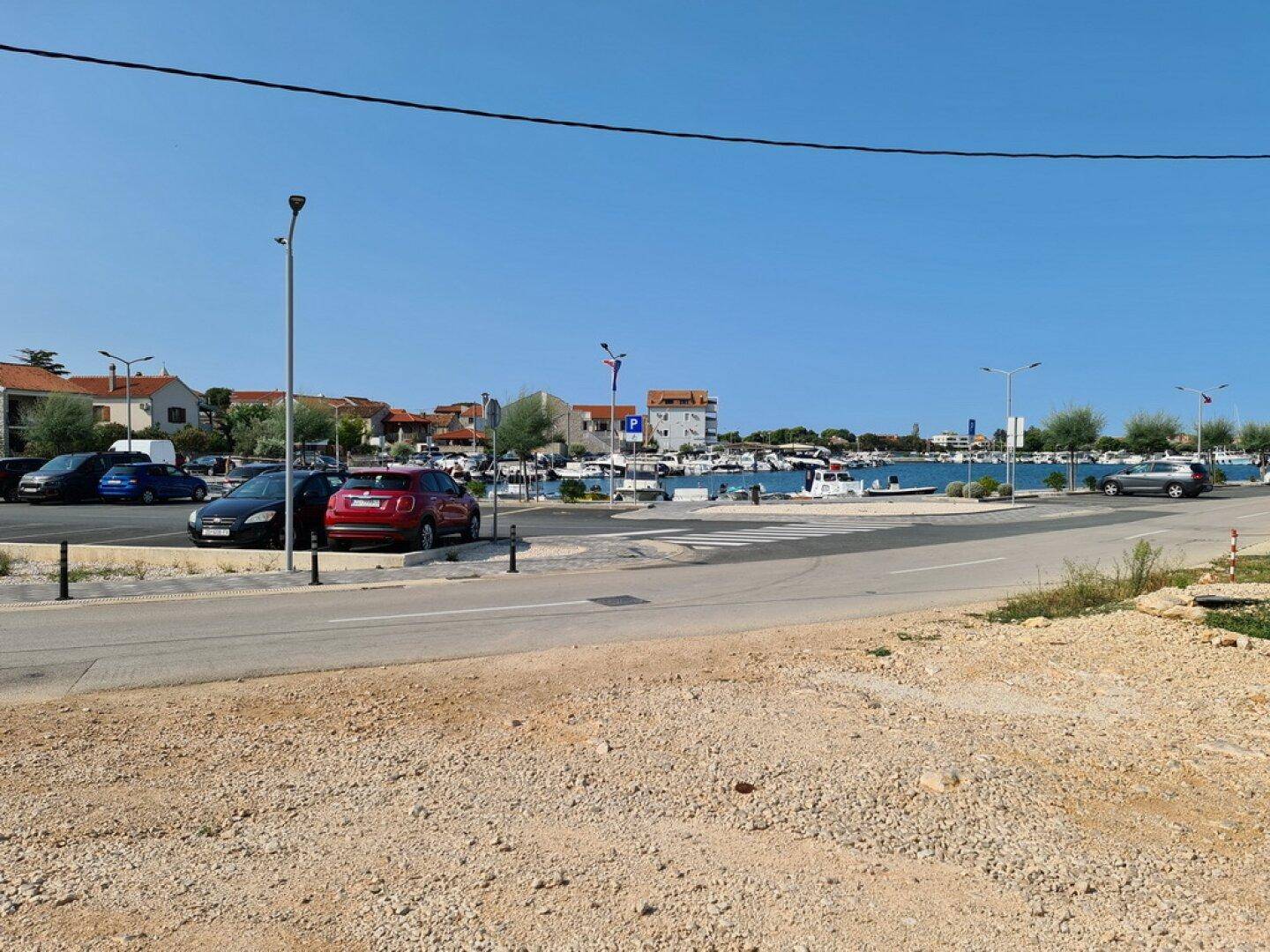 Blick auf eine Küstenstraße mit parkenden Autos und einem Hafen im Hintergrund unter blauem Himmel.