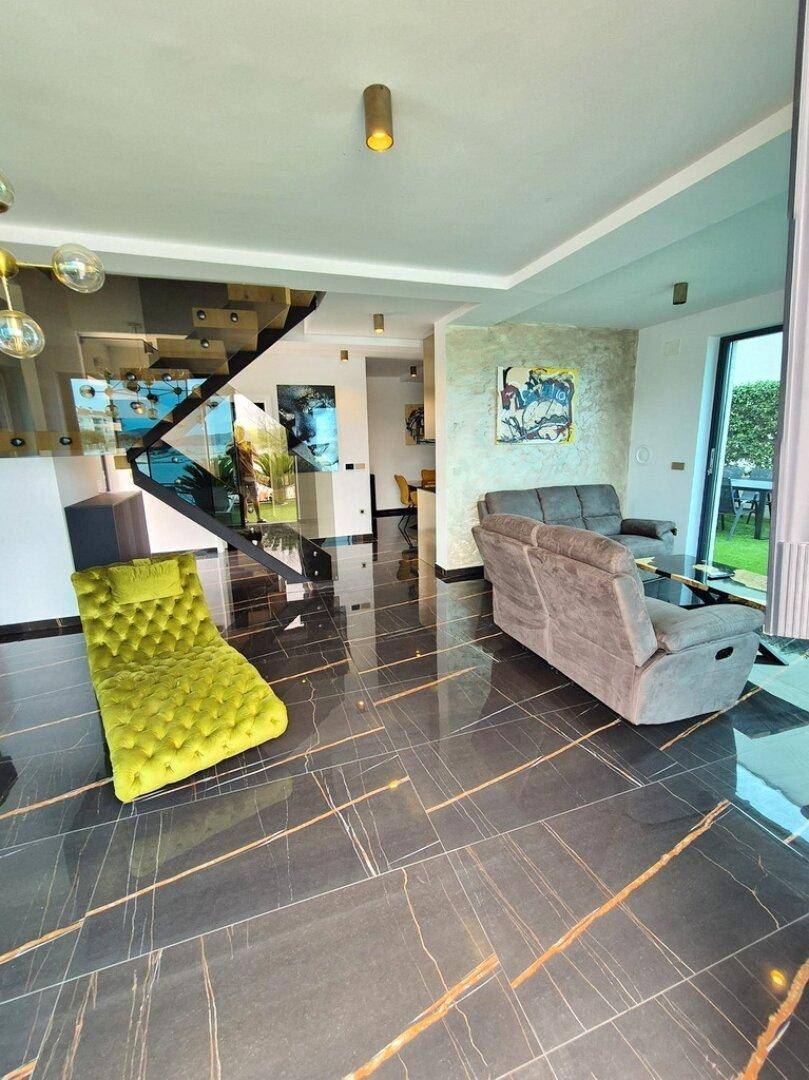 Elegantes Wohnzimmer mit glänzenden dunklen Fliesen, moderner Treppe und Blick ins Grüne.