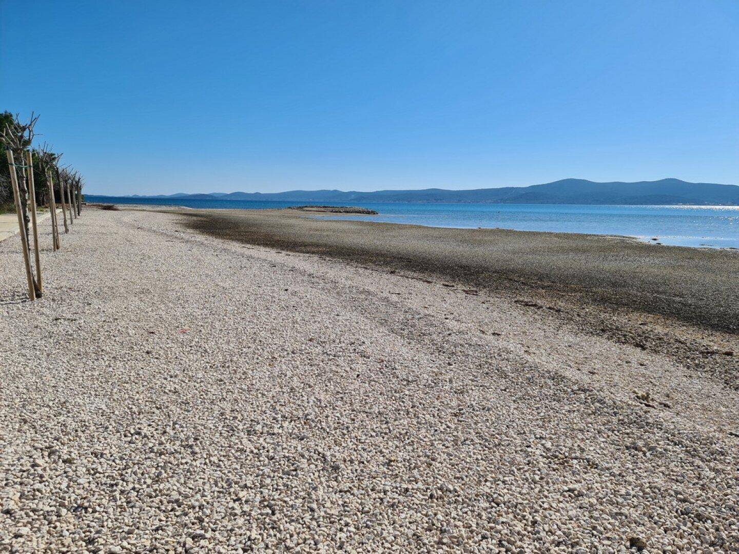 Weiter Kiesstrand mit Blick auf das ruhige Meer und die entfernten Berge unter blauem Himmel.