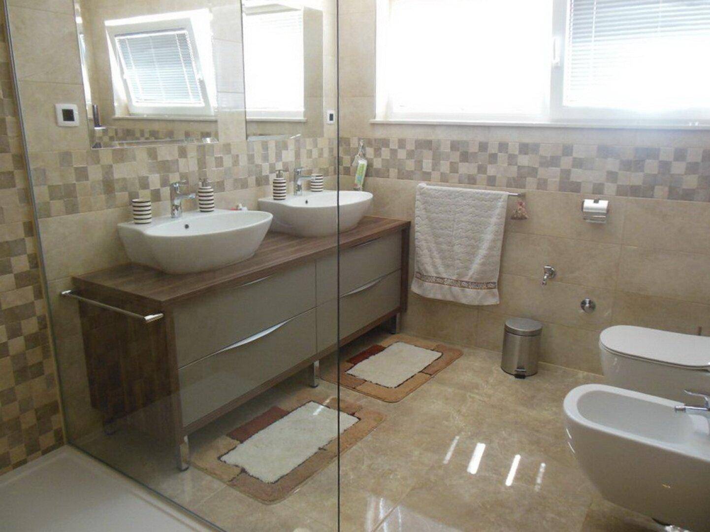 Modernes Badezimmer mit Doppelwaschbecken, Bidet, Dusche und beigen Fliesen sowie Fenster.