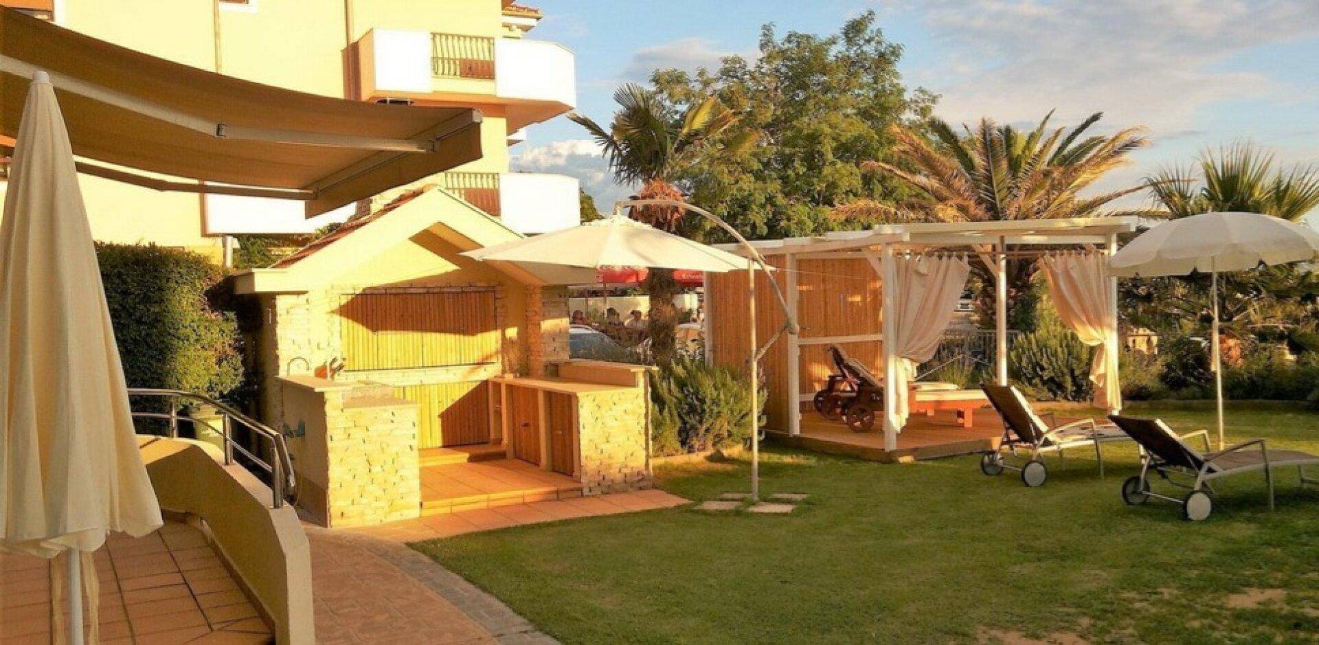 Garten mit Außenküche, schattiger Cabana, Sonnenliegen und üppiger mediterraner Vegetation.