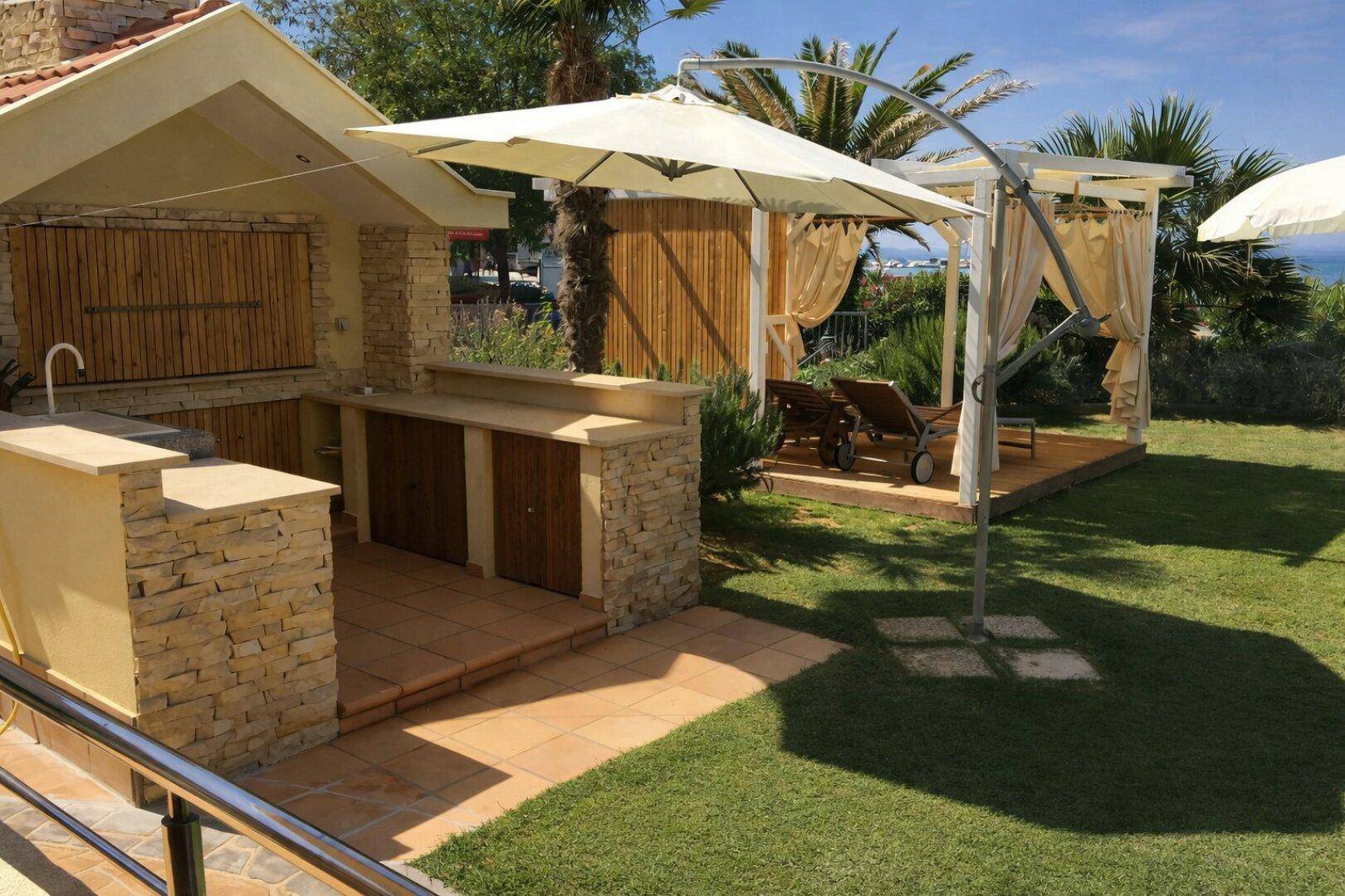 Gartenbereich mit Außenküche, Rasenfläche, Sonnenliegen und einer schattigen Cabana unter Palmen.