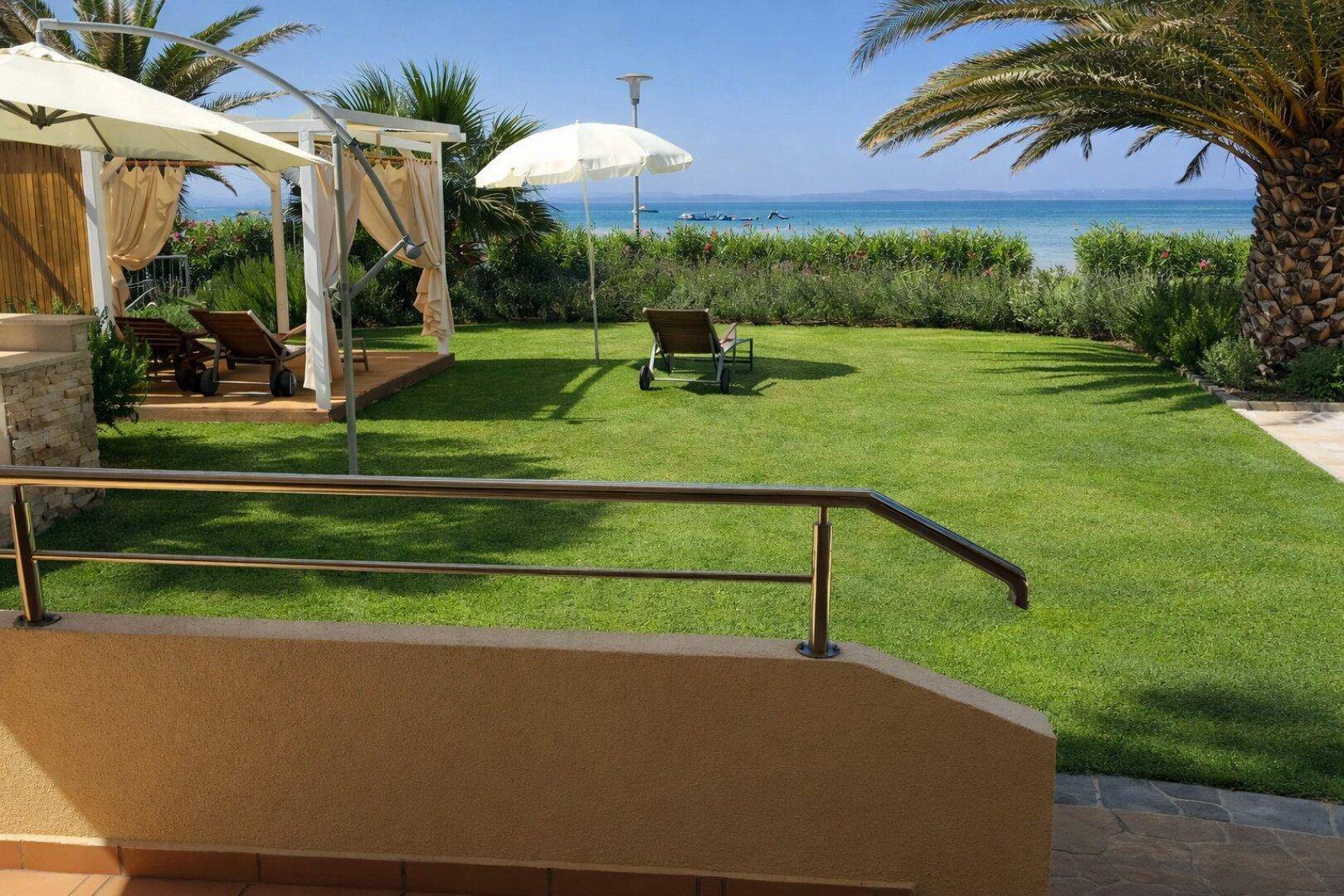 Grüner Garten mit Rasenfläche, Sonnenliegen, Cabana und weitem Blick auf das blaue Meer.