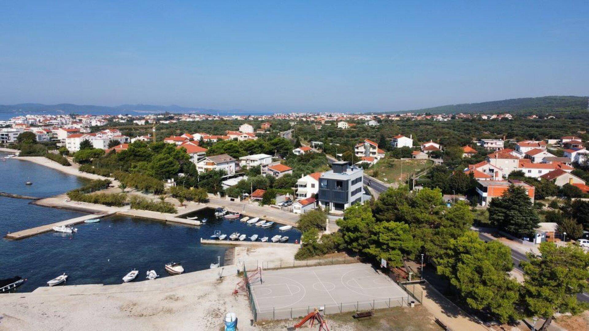 Weitläufige Luftaufnahme einer Küstenlandschaft mit Wohnbebauung, Meer und Sportplatz.
