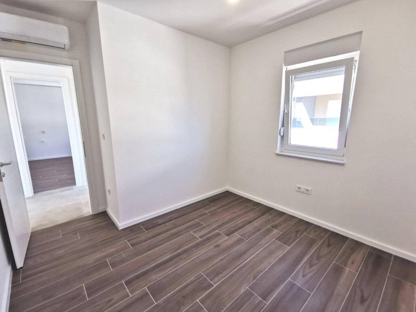 Unmöbliertes Schlafzimmer mit modernem Fliesenboden in Holzoptik und Fenster, bietet viel Tageslicht.