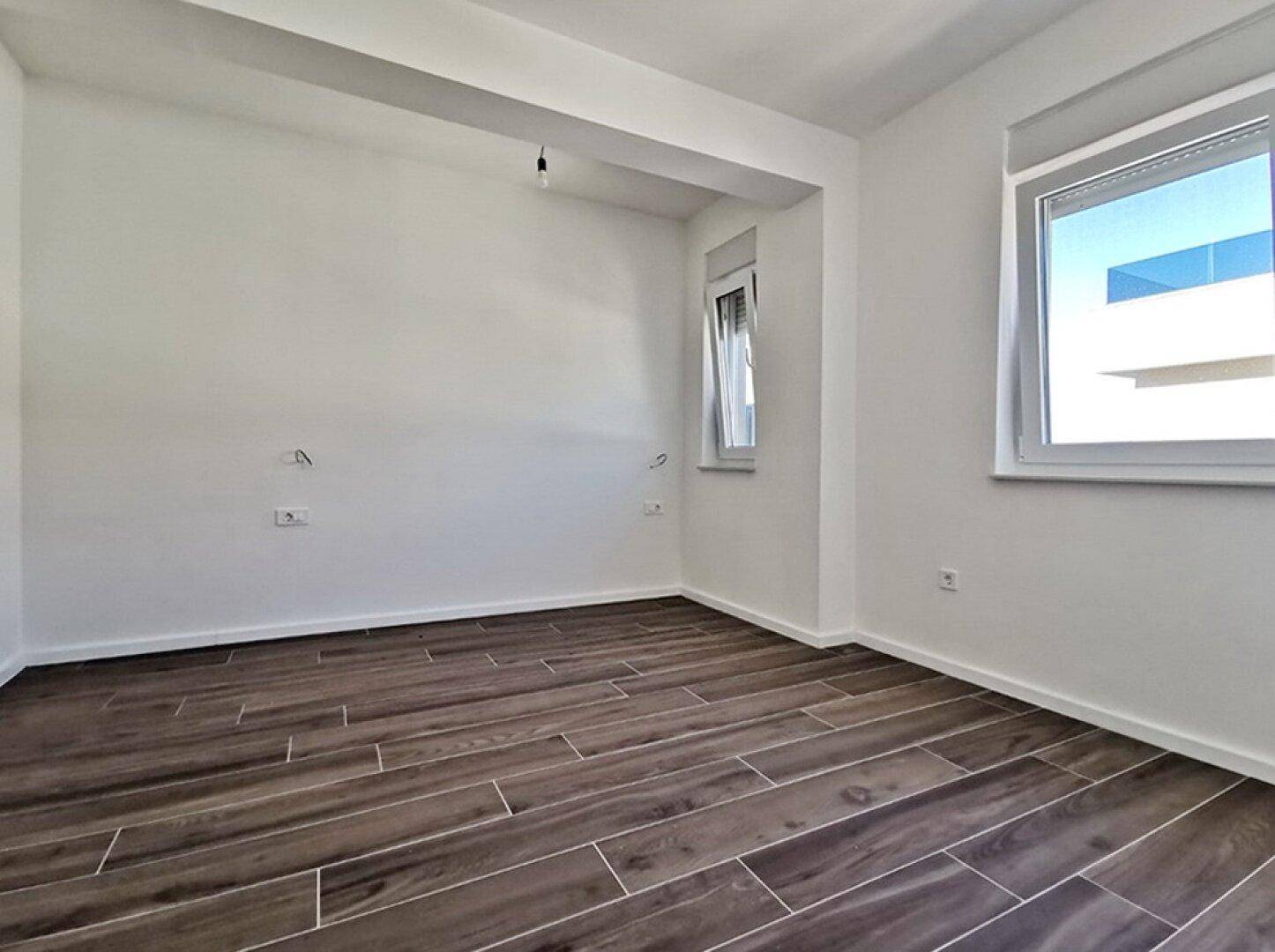 Geräumiges Schlafzimmer mit Fliesenboden in Holzoptik und zwei Fenstern, bietet viel Helligkeit.