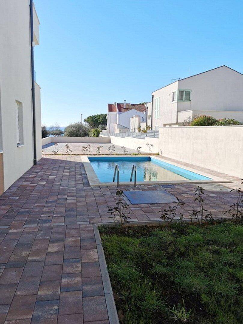 Außenbereich mit kleinem Pool und gepflasterter Fläche, umgeben von jungen Pflanzen und Gebäuden.