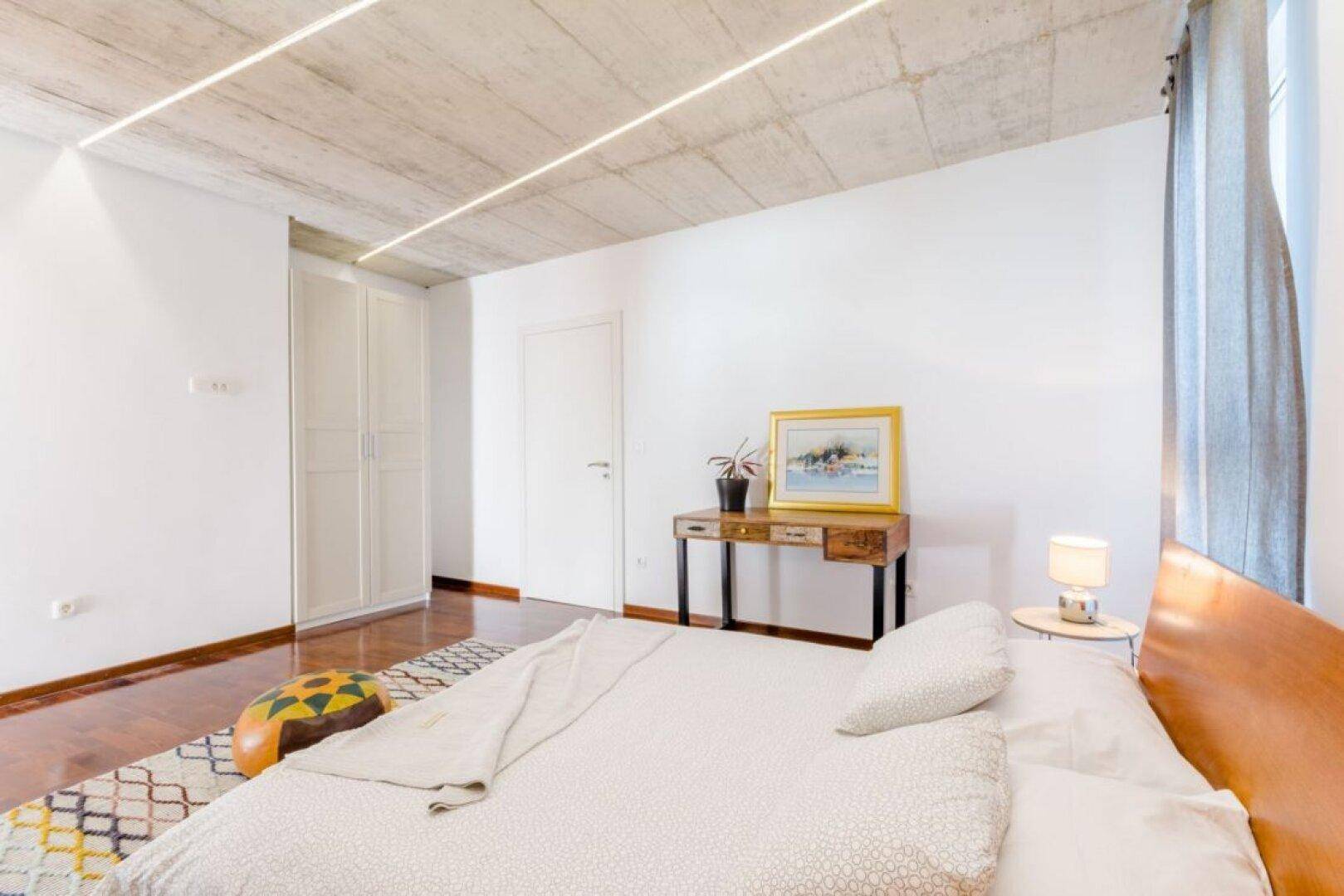 Helles Schlafzimmer mit Holzboden, weißem Kleiderschrank und sichtbarer Betondecke.
