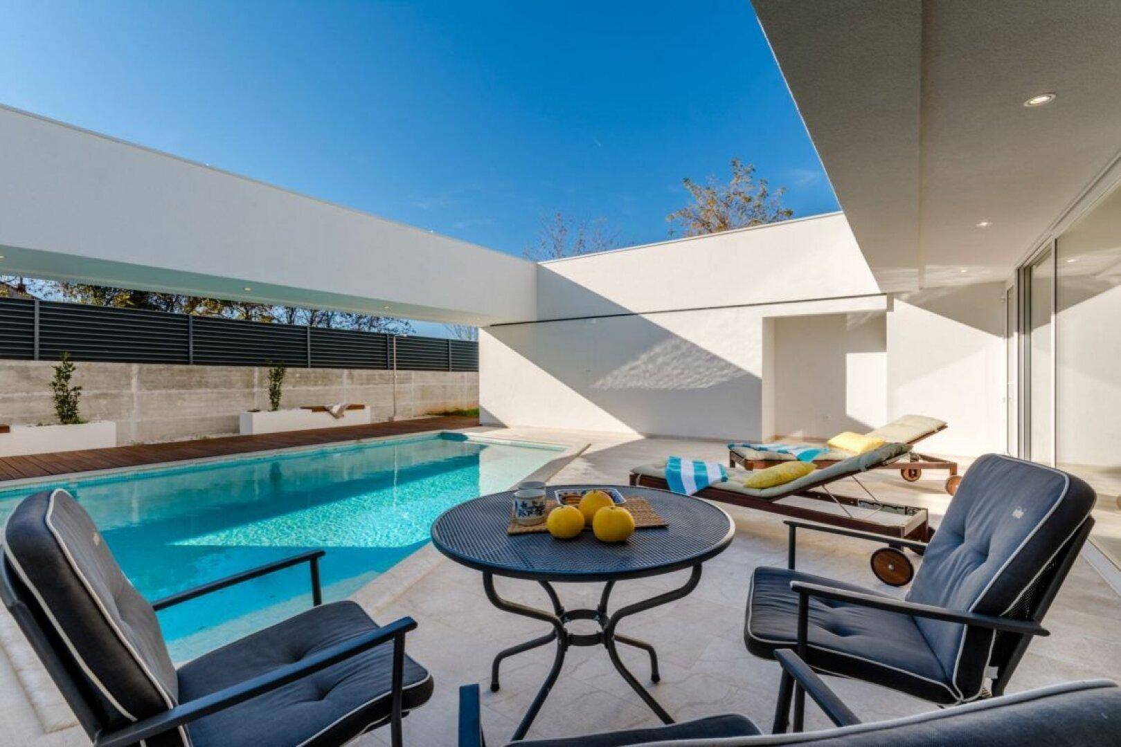 Gemütlicher Sitzbereich am Pool mit bequemen Stühlen und einem kleinen Tisch unter blauem Himmel.
