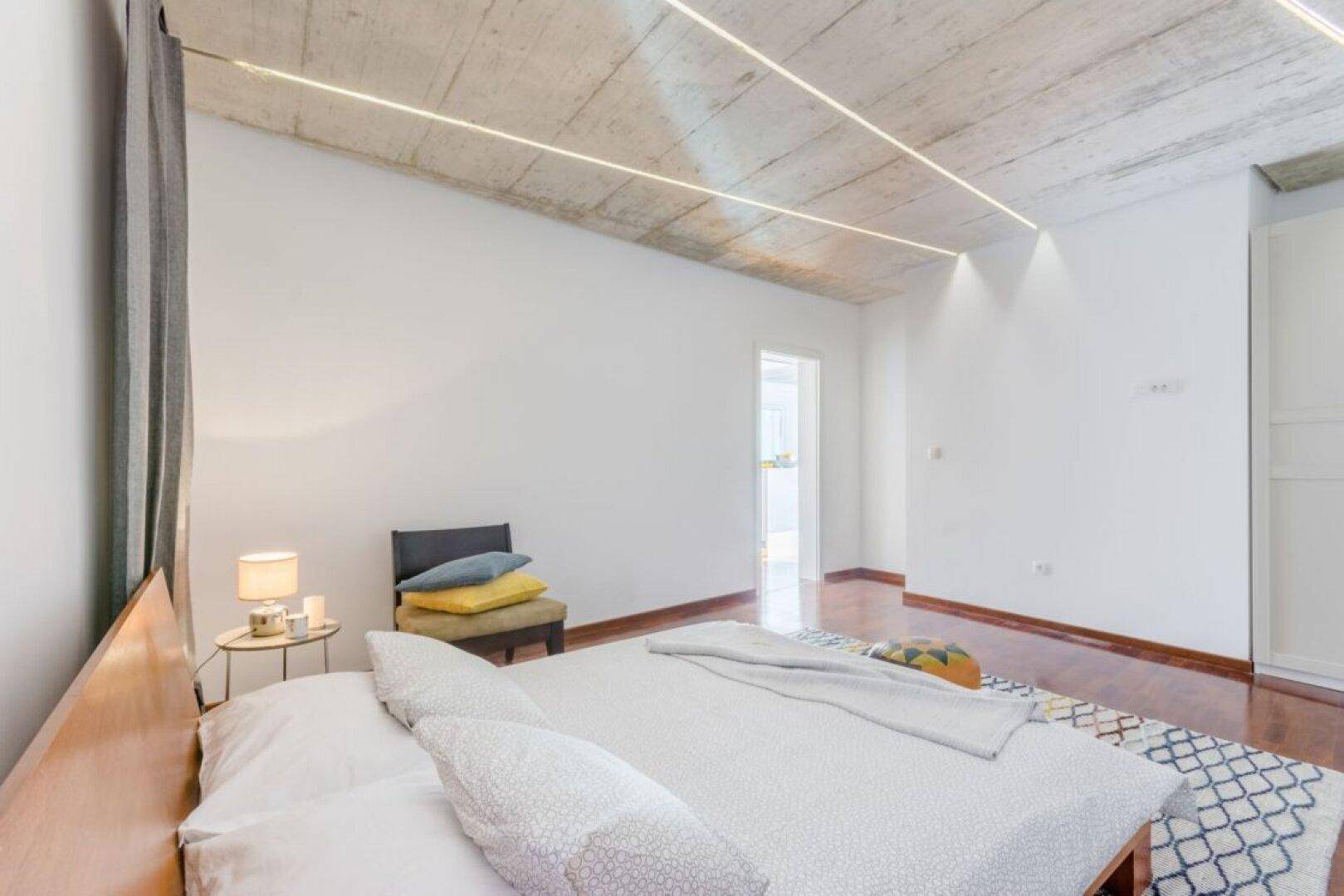 Schlafzimmer mit Holzboden, schlichtem Bett und sichtbarer Betondecke, durchflutet von natürlichem Licht.