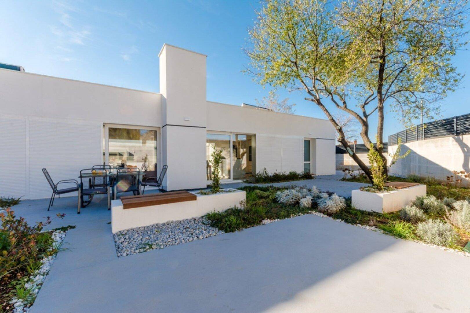 Rückseite der modernen Villa mit gepflastertem Patio, Sitzgelegenheiten und mediterraner Bepflanzung.