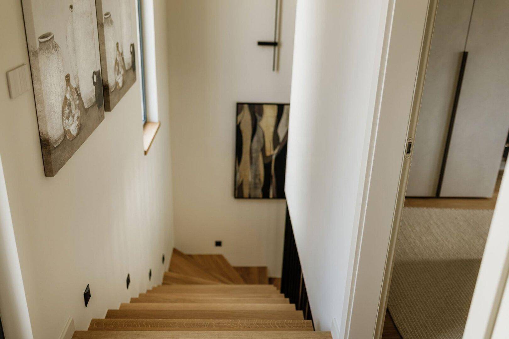Holztreppe führt nach unten, beleuchtet von Wandleuchten und natürlichem Licht.