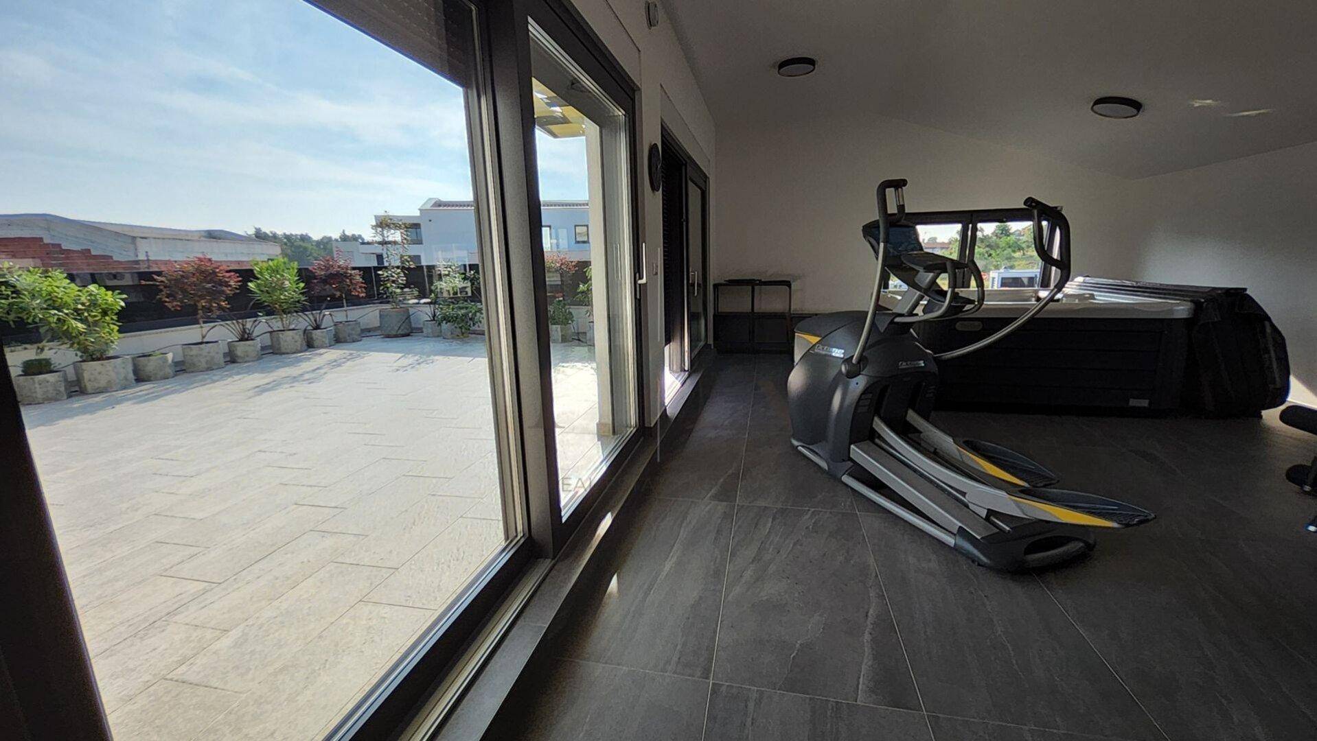 Heller Fitnessbereich mit Trainingsgeräten und weitem Blick auf die großzügige Dachterrasse.